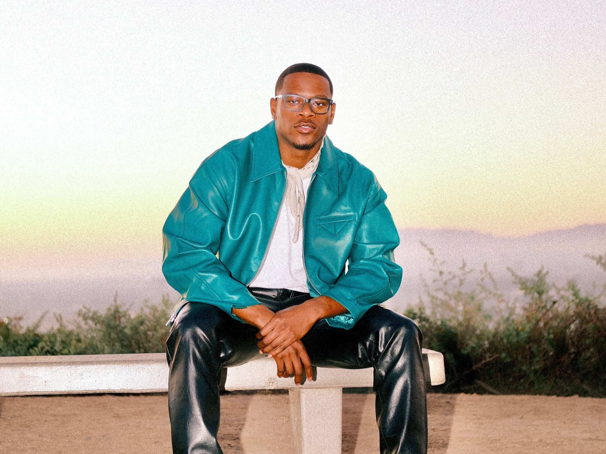 Young man in teal jacket sitting outdoors against a sunset sky.