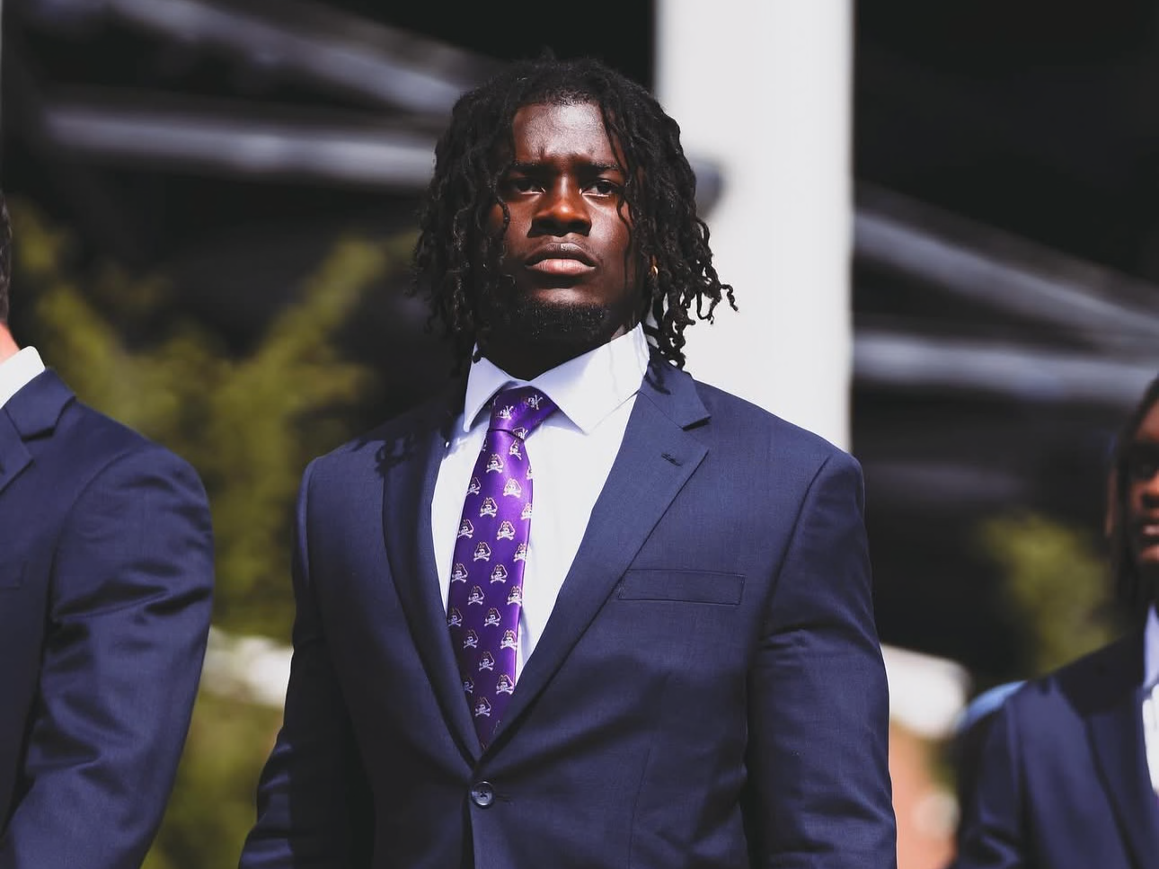 Man in a navy suit with a purple tie standing outdoors.