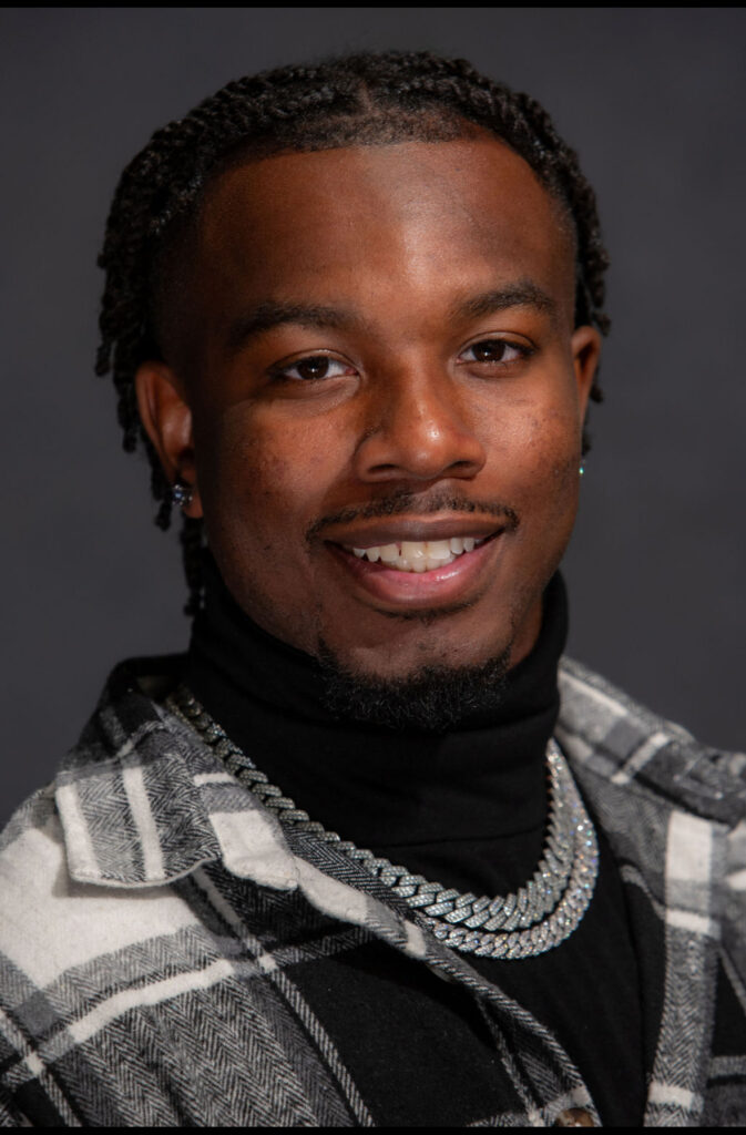 A smiling man wearing a checkered jacket and chain necklaces.