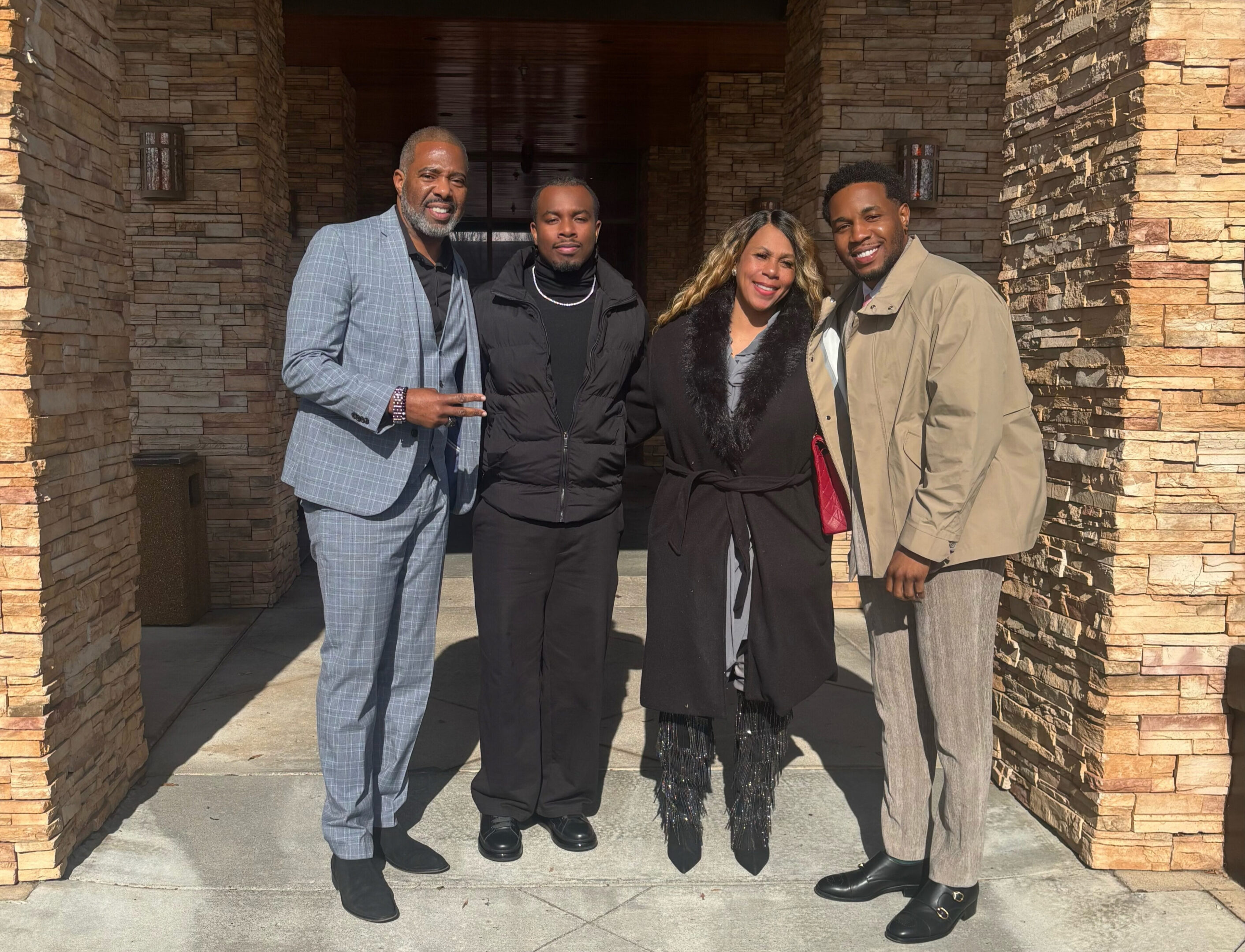 Four people posing together outside a stone building on a sunny day.