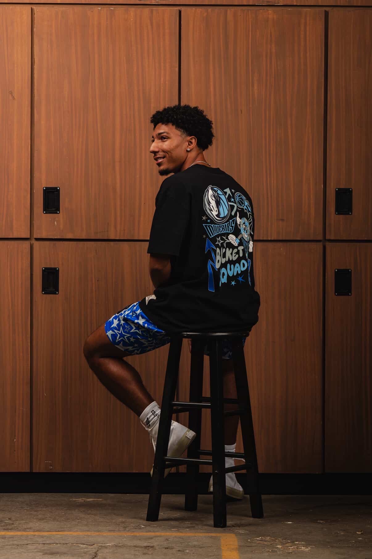 Young man sitting on a stool wearing a blue backpack and patterned shorts.